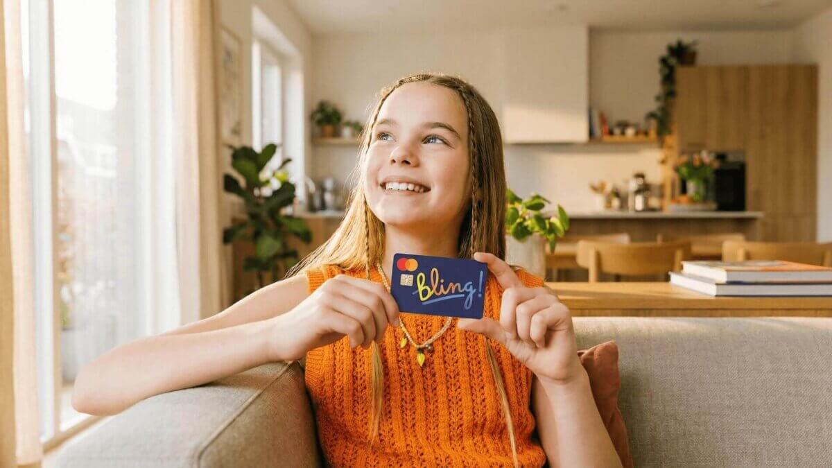 Ein Mädchen mit langen Haaren und orangefarbenem Pullover hält die charakteristische dunkelblaue Bling-Karte mit dem bunten „bling!"-Schriftzug und Mastercard-Logo – in einem hellen, modernen Wohnzimmer mit warmem Tageslicht.