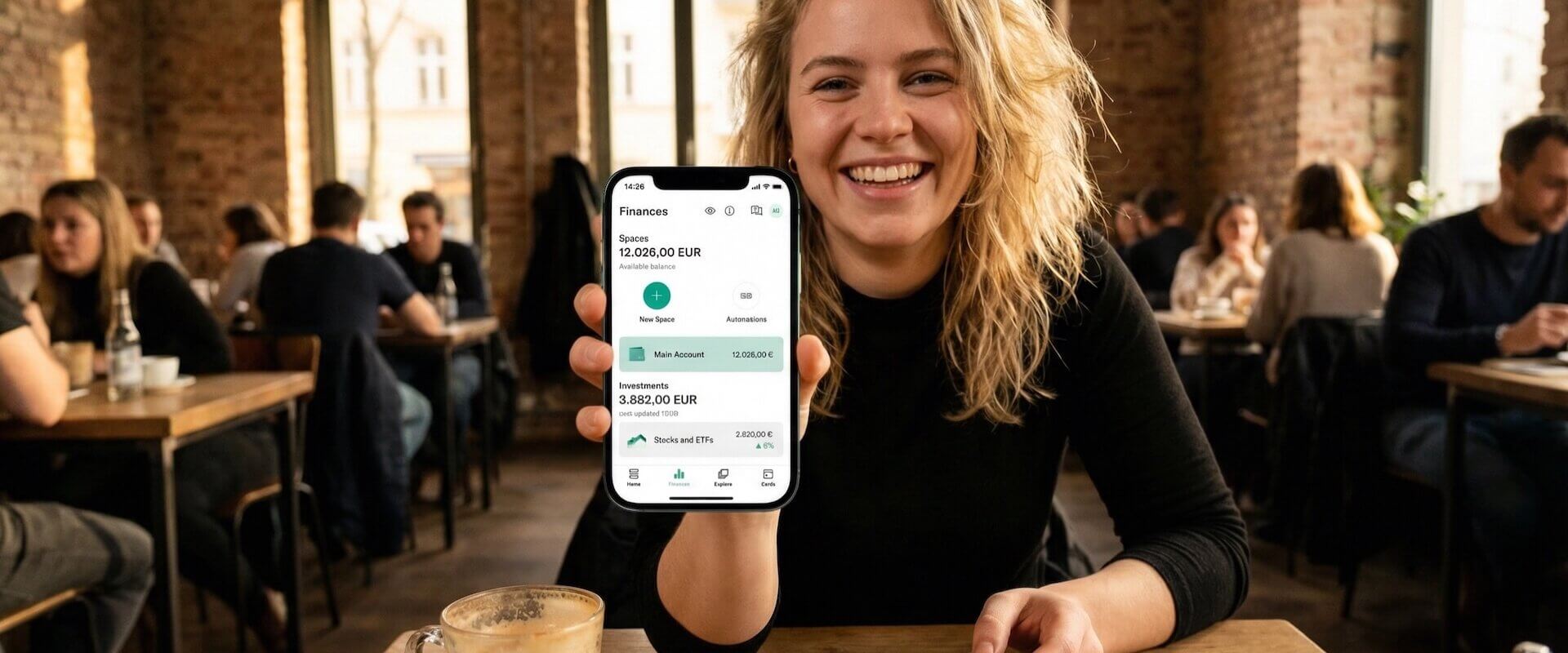 Junge Frau im Berliner Café zeigt N26 App mit Finances-Übersicht, 12.026 Euro Kontostand und 3.882 Euro Investments in Aktien und ETFs, transparente N26-Debitkarte in der Hand