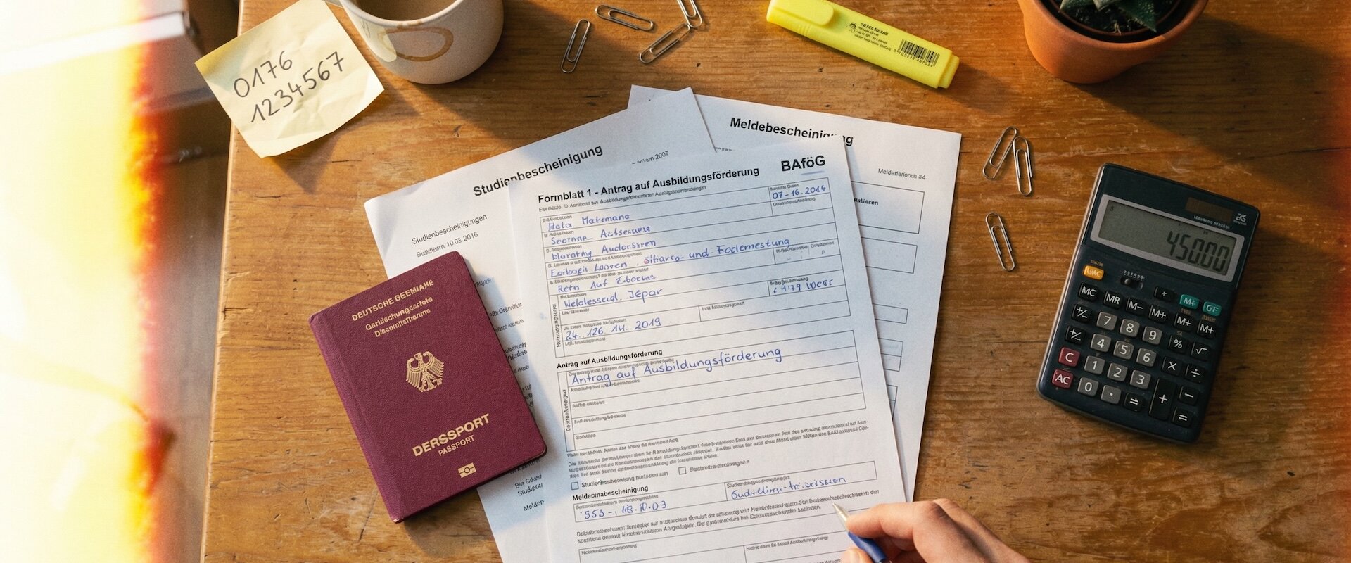 BAföG-Antragsformular auf Holzschreibtisch mit Reisepass, Studienbescheinigung, Taschenrechner und Kaffeetasse, Hand hält Kugelschreiber, warmes Filmkorn