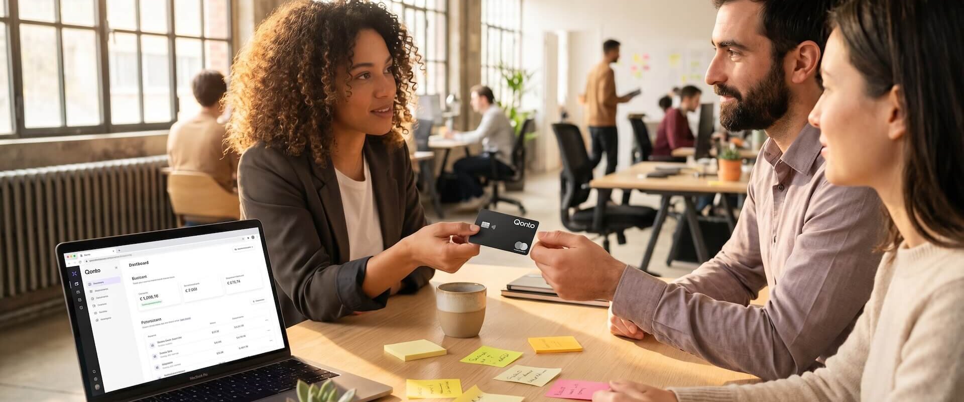 Diverses Business-Team im modernen Co-Working-Büro reicht eine schwarze Qonto Mastercard weiter, Laptop zeigt das Qonto-Dashboard