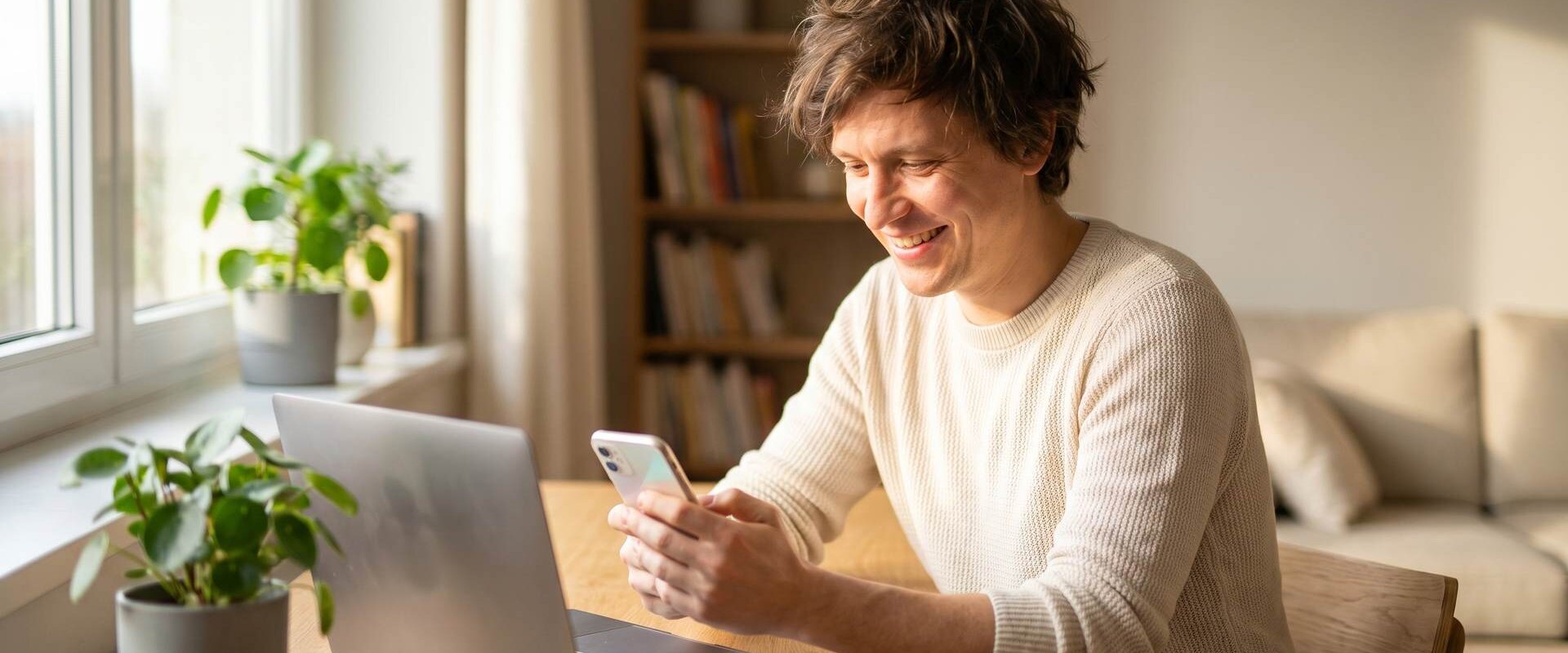 Lächelnder Mann beim Online-Banking mit dem Smartphone an einem hellen Schreibtisch zu Hause